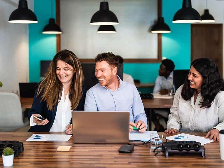 Three professionals sitting at a desk with an open laptop working at a shared co-working space and smiling as woman holds up phone to share information with the other two people.